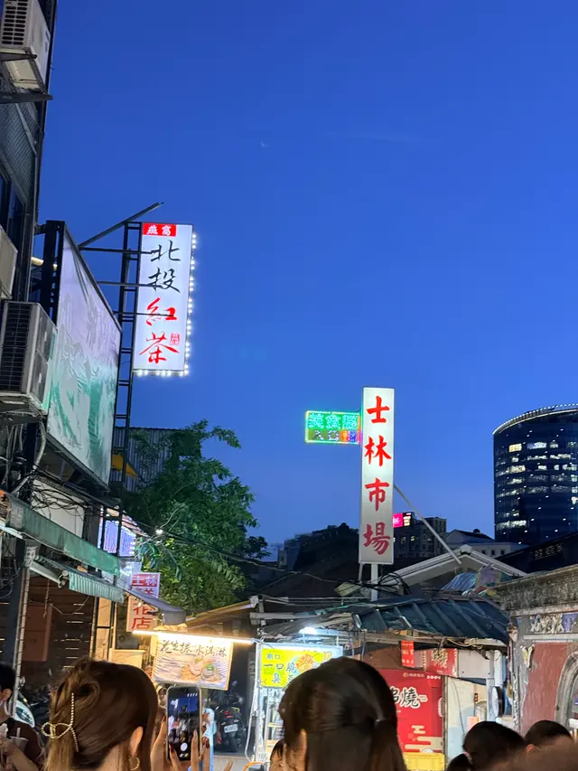 Photographed at Shilin Night Market in Taipei, where the vertical market sign glows beneath a blue-hour sky and stall lights lead into the crowded old street.