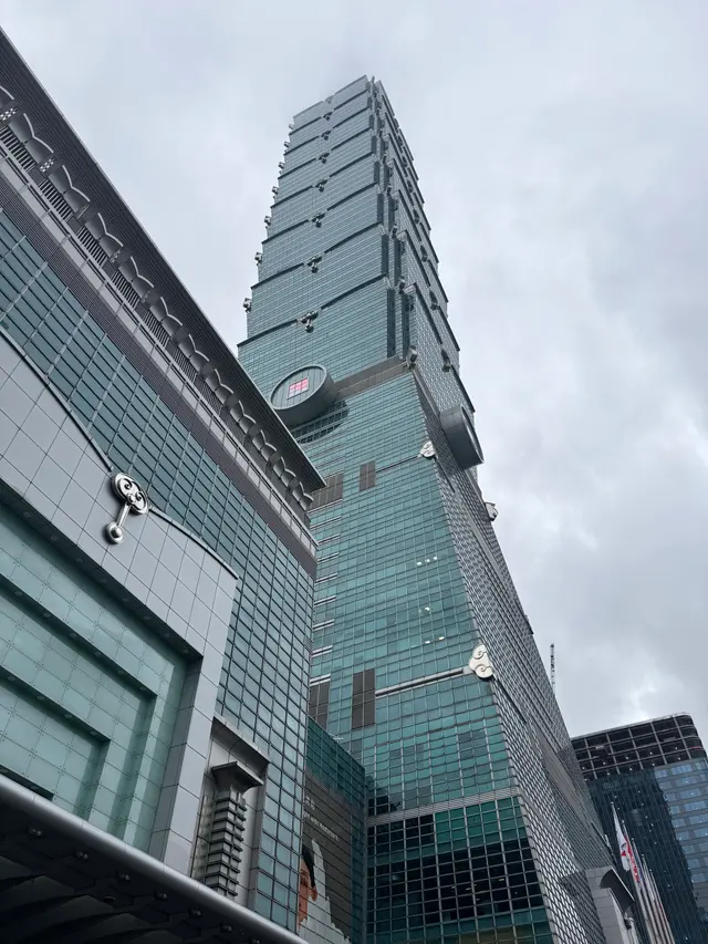 Photographed in Taipei's Xinyi District, looking up at Taipei 101 as its layered glass facade and projecting forms stand sharply against an even gray sky.