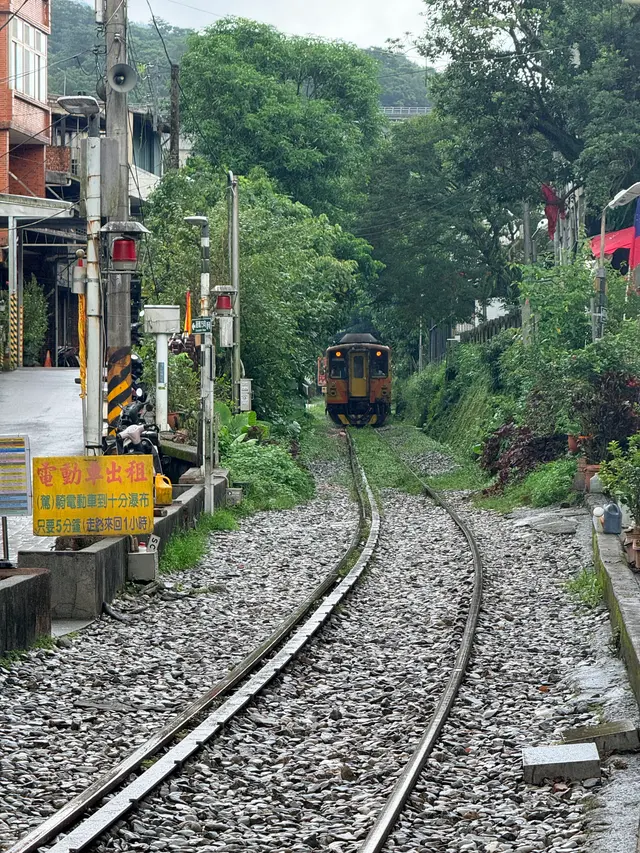 Photographed on Shifen Old Street in New Taipei, where a train approaches along rain-darkened tracks between greenery and homes as warning lights glow beside the shops.