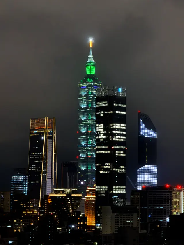Photographed from Songshan in Taipei, where Taipei 101 glows green above the Xinyi skyline beneath a damp, low night sky.