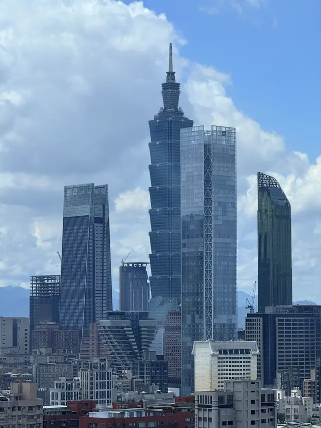 Photographed from above in Taipei's Songshan district, looking across the city toward Taipei 101 as the Xinyi skyline rises beneath summer clouds and blue sky.