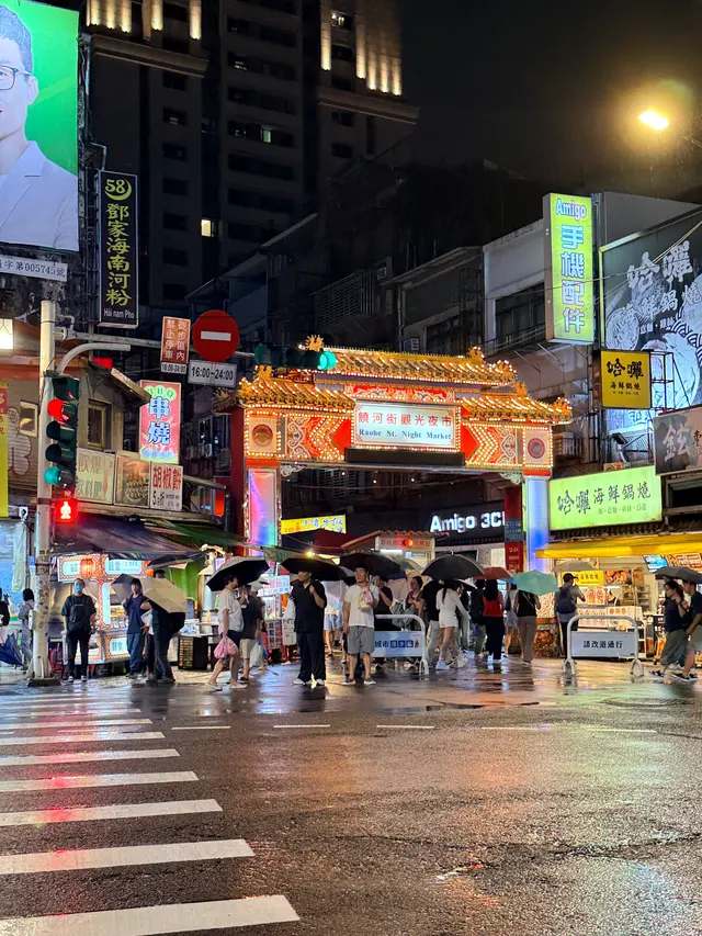 Photographed at Raohe Street Night Market in Taipei, where crowds under umbrellas pass beneath the illuminated gate and wet pavement reflects the colors of stalls and signs.