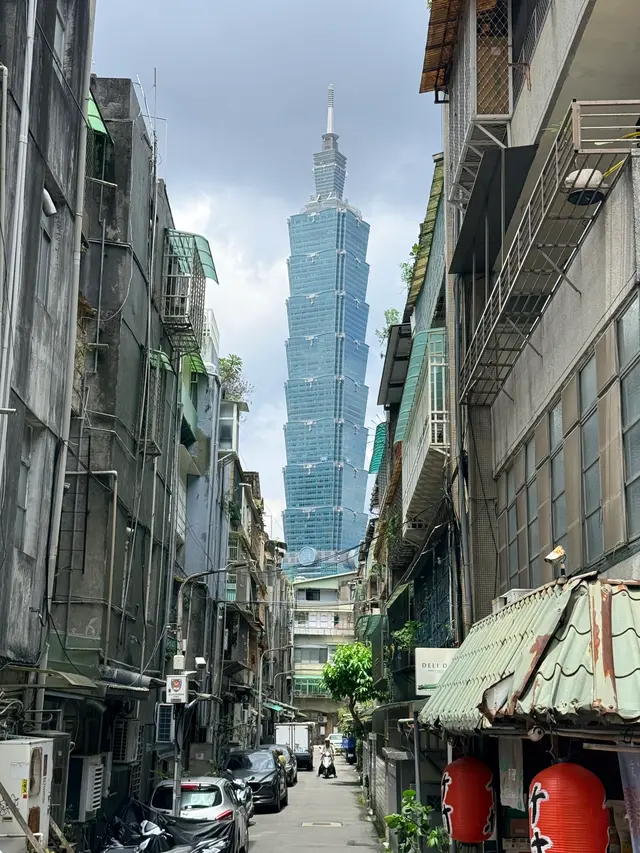 Photographed in Taipei's Xinyi District, where a narrow lane framed by aging apartments and metal window grilles opens directly toward Taipei 101, layering old and new city scales beneath a clear sky.