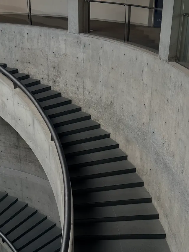 Photographed at He Art Museum in Foshan, where dark steps sweep along exposed concrete walls and a curved handrail links the levels in one fluid line.