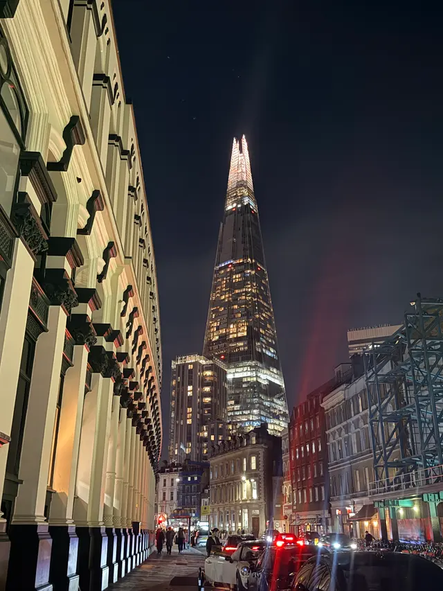 In London, the Shard's illuminated spire rises above the street, where a colonnade recedes into the distance and headlights mingle with warm lights along the pavement.